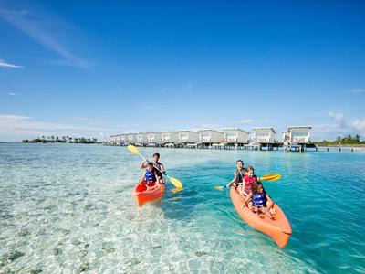 Twee personen kajakken op helder turquoise water voor een resort met bungalows.