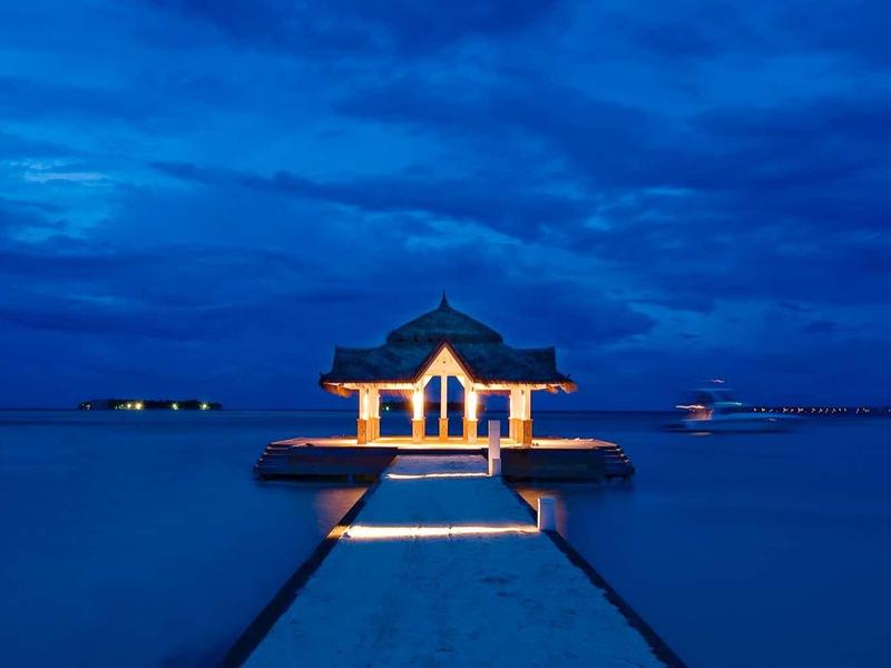 Ein beleuchteter Pavillon am Ende eines Stegs über ruhigem Wasser bei blauem Nachthimmel.