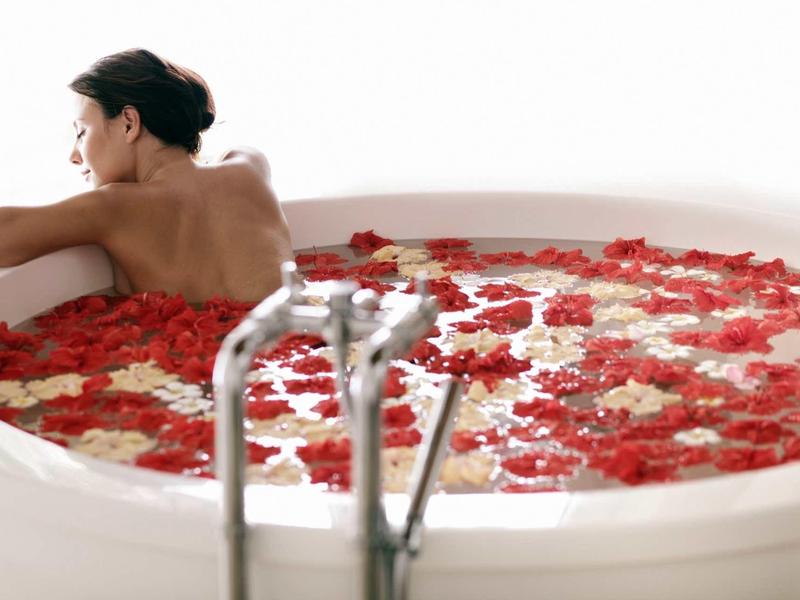 Frau entspannt in weißer Badewanne mit Wasser und roten sowie weißen Rosenblättern.