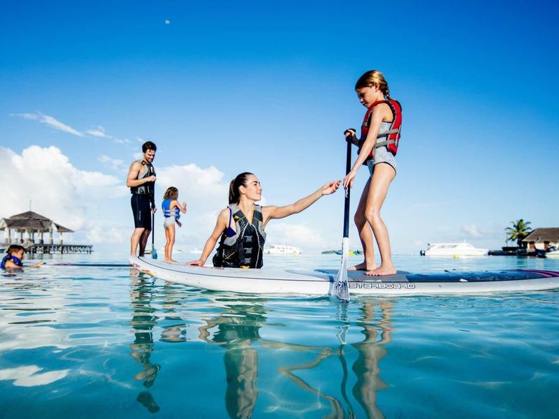 Vier Personen mit Schwimmwesten stehen und sitzen auf einem Paddleboard im klaren blauen Wasser.