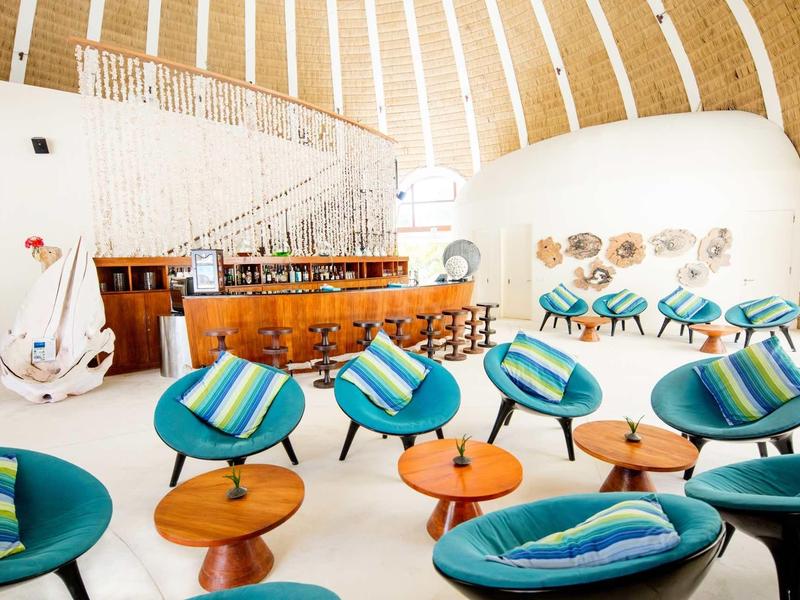 Moderne Lounge mit blauen Sesseln, Holztischen, Bar und geschwungener Treppe in heller, luftiger Halle.