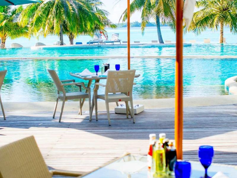 Freiluftrestaurant mit Tischen, blauen Gläsern, Sonnenschirmen und Pool mit Palmen im Hintergrund.