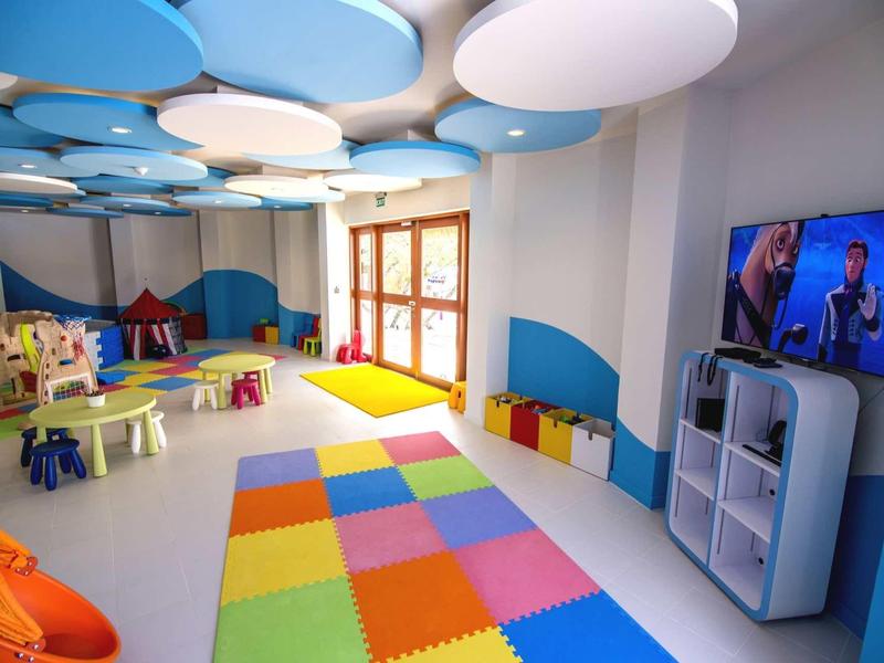 Bunter Kindergartenraum mit Spielzeug, bunten Matten, kleinen Tischen und Stühlen, großer Fensterfront und TV.