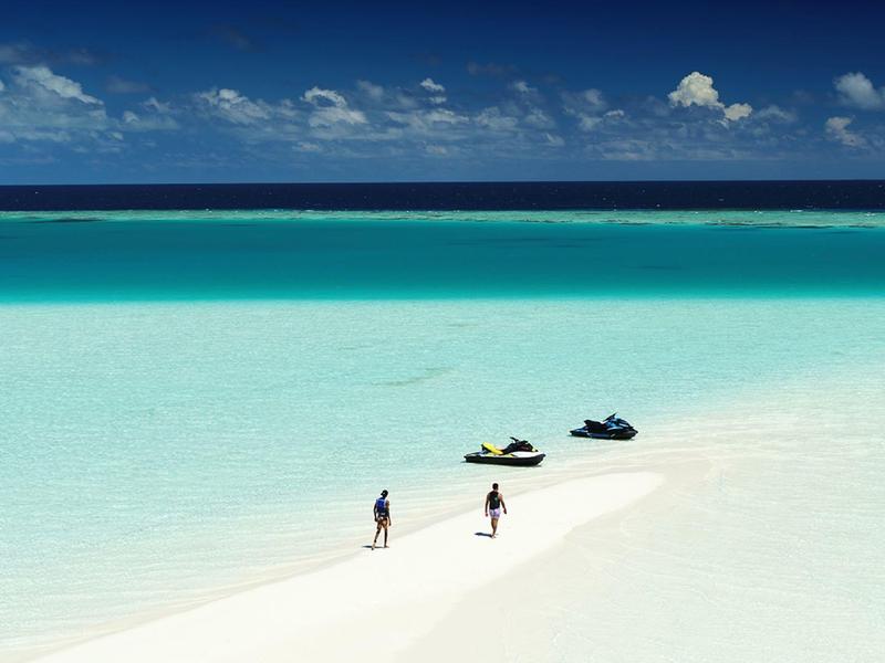 Zwei Personen laufen auf schmalem Sandweg im kristallklaren türkisblauen Meer bei sonnigem Himmel.