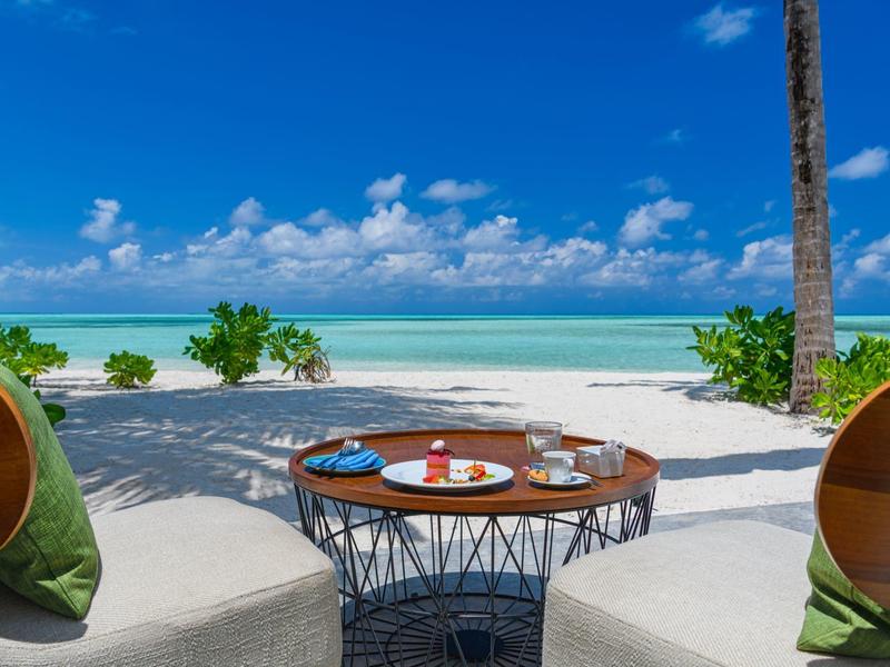 Strand mit weißem Sand, blauem Meer und wolkigem Himmel, zwei bequeme Sessel und ein Tisch mit Frühstück.