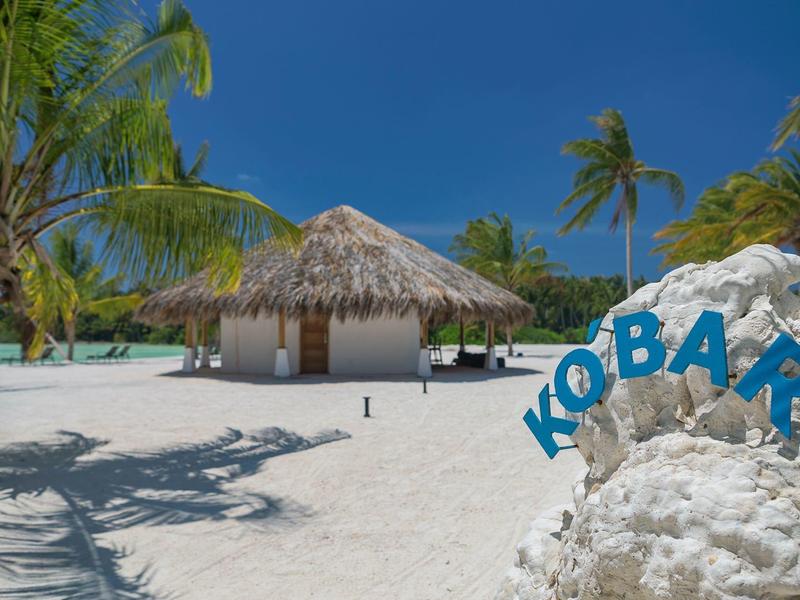 Strand mit weißen Sand, Palmen, einer Strohhütte und blauer Schrift "KOBAR" auf Felsen.