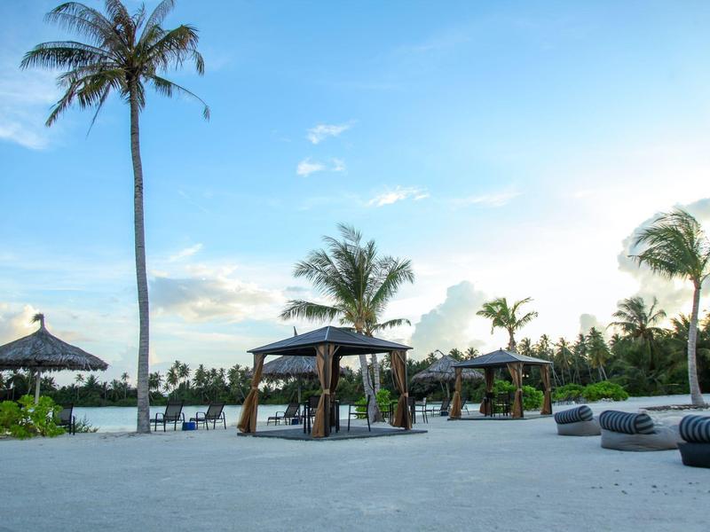 Badebereich mit Sonnenliegen, Pavillons und Palmen unter blauem Himmel am Wasser.