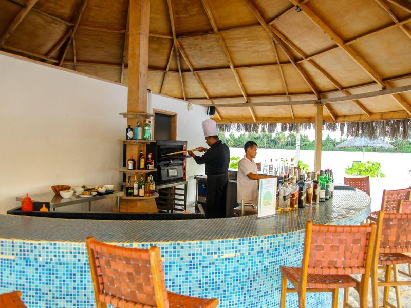 Offene Bar mit Mosaikfront, Holzdach und mehreren Hockern am Strand mit Blick auf Palmen.