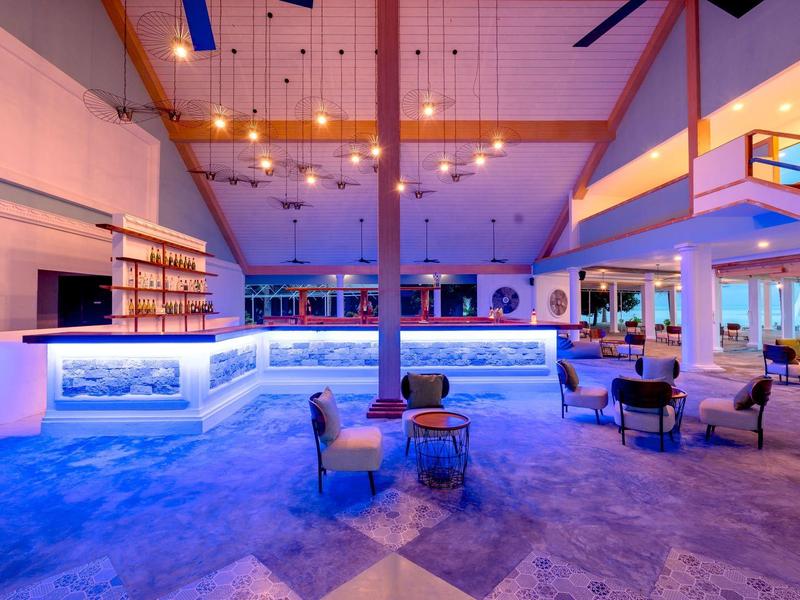 Großer moderner Raum mit beleuchteter Bar, gemütlichen Sesseln und Holzdecken mit Pendelleuchten.