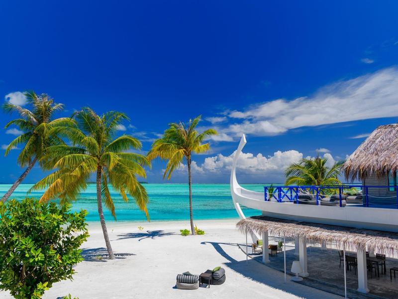 Strand mit weißem Sand, Palmen, blauem Himmel, türkisfarbenem Meer und einem Holzhaus mit Strohdach.