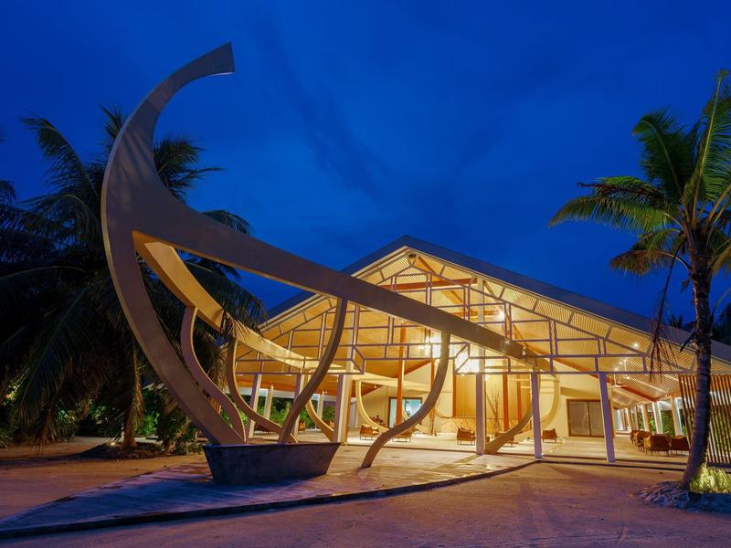 Modernes Gebäude mit geschwungenen weißen Strukturen, großen Glasflächen und Palmen bei Abenddämmerung.