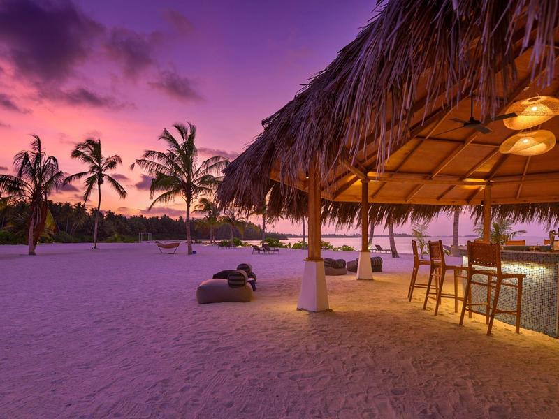 Strandbar mit Strohdach, Holztheke und Sitzplätzen am Sandstrand bei lila Sonnenuntergang.