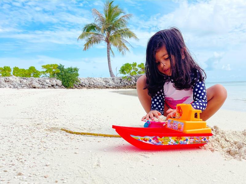 Ein Mädchen spielt mit einem roten Spielzeugboot am weißen Sandstrand unter blauem Himmel.