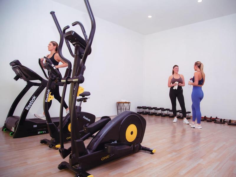 Zwei Frauen trainieren im hellen Fitnessraum mit Crosstrainer, Laufband und Hanteln.