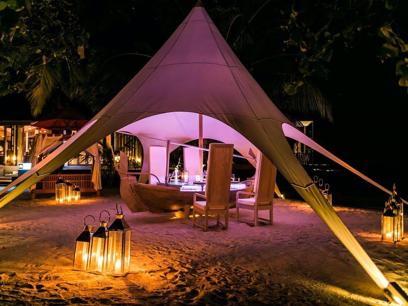 Beleuchtetes weißes Zelt mit Sitzmöbeln und Laternen auf sandigem Boden bei Nacht.