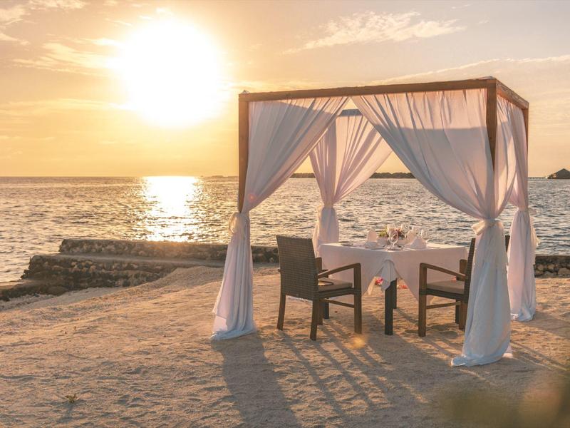 Ein Tisch mit zwei Stühlen unter einem Baldachin am Strand bei Sonnenuntergang.