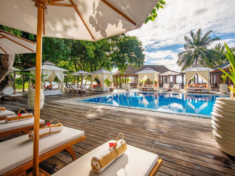 Poolbereich mit Liegestühlen, Sonnenschirmen, Palmen und Holzdeck unter blauem Himmel.