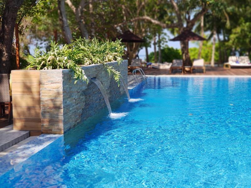 Ein klarer blauer Pool mit Wasserfällen und Sonnenschirmen im Hintergrund unter Bäumen.