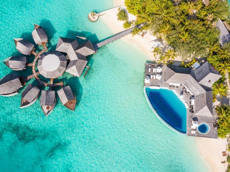 Überwasser-Bungalows auf türkisfarbenem Wasser neben weißem Sandstrand und Poolbereich.