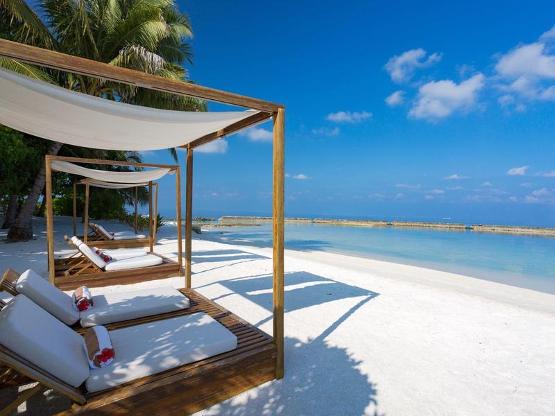 Strand mit weißen Liegen unter Sonnensegeln, Palmen und klarem blauem Meer bei sonnigem Himmel.