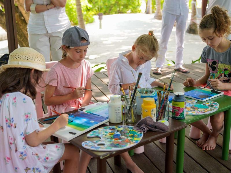 Kinder malen konzentriert an einem Tisch im Freien, umgeben von Farben und Pinseln.