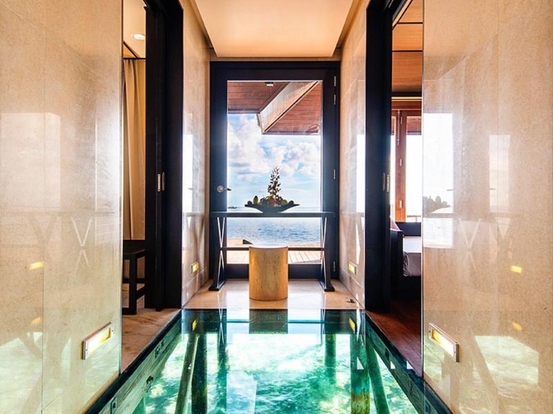 Flur mit glänzendem, grünem Glasboden, seitlichen Türen und Aussicht auf Wasser und Statue draußen.