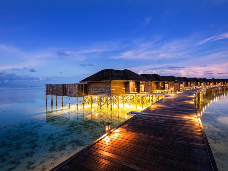 Ein heller Steg führt zu über dem Wasser gebauten Hütten bei Sonnenuntergang.