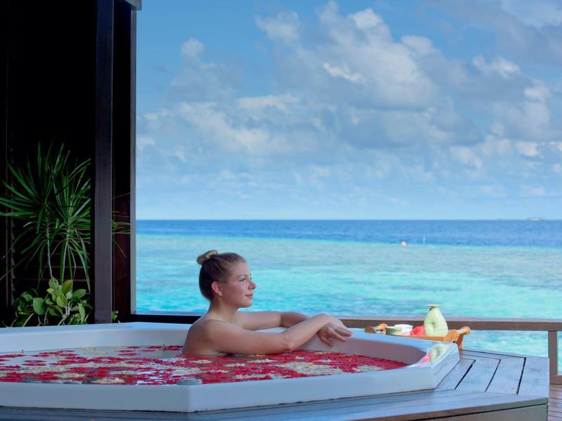 Frau entspannt in runder Badewanne mit Blumen, Meer und Himmel im Hintergrund sichtbar.
