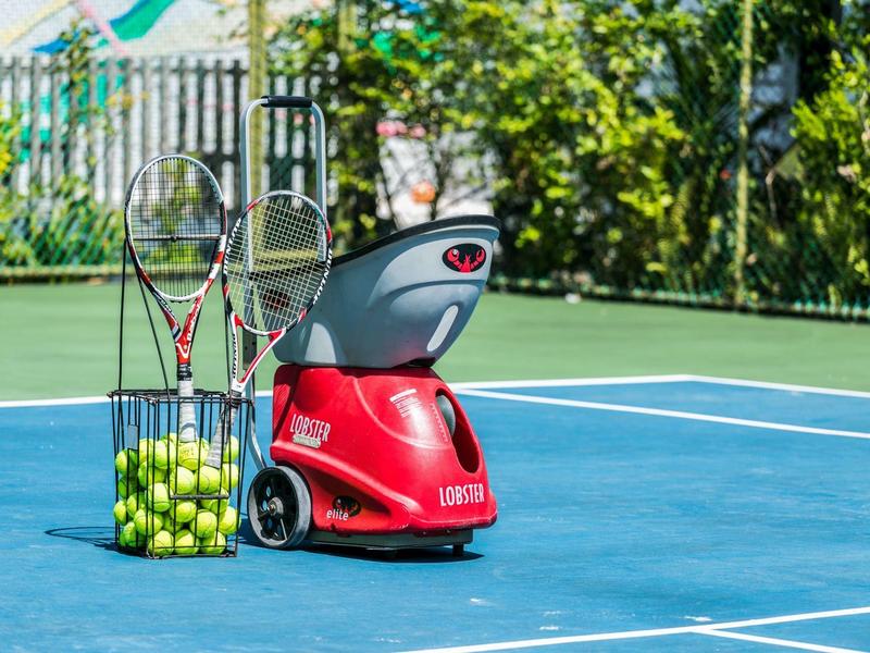 Rote Tennisballmaschine auf blauem Tennisplatz neben Korb mit Schlägern und vielen gelben Bällen.