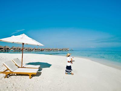 Frau steht am tropischen Strand neben Liegestühlen und Sonnenschirm bei klarem Himmel.
