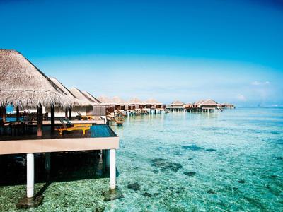 Reihenbungalows mit Strohdächern auf Stelzen über klarem, türkisfarbenem Wasser und blauem Himmel.