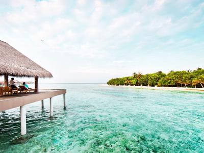 Ein Steg mit Strohdach über klarem, türkisfarbenem Wasser, mit Palmen am Strand im Hintergrund.