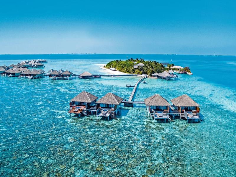 Bungalows auf Stelzen über klarem Wasser nahe einer tropischen Insel mit blauem Himmel.