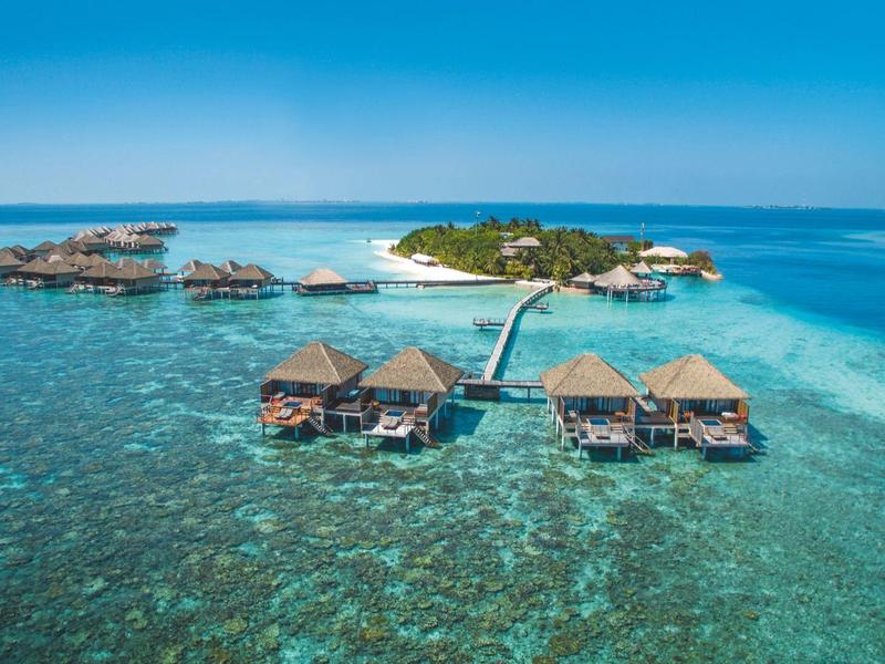 Bungalows auf Stelzen über klarem, türkisfarbenem Wasser mit kleinem Inselhintergrund und Steg.
