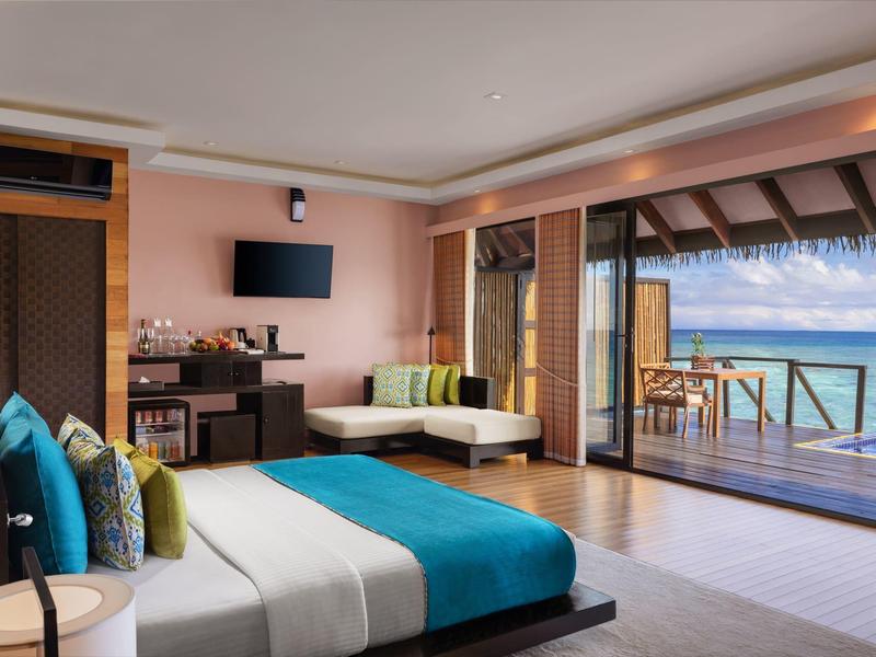 Modernes Hotelzimmer mit großem Bett, Sitzbereich und Holzterrasse mit Meerblick bei Sonnenschein.