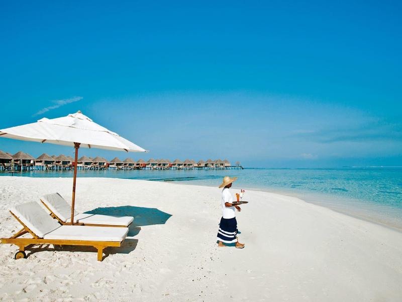 Frau steht am tropischen Strand neben Liegestühlen und Sonnenschirm bei klarem Himmel.