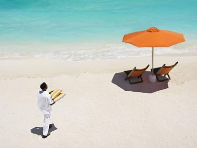 Un serveur portant des plateaux marche sur la plage sablonneuse vers deux chaises longues et un parasol.