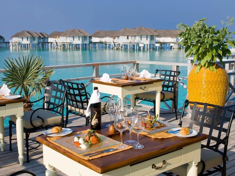 Eine Terrasse mit Tischen und Stühlen, Meerblick und Häuser am Wasser bei blauem Himmel.