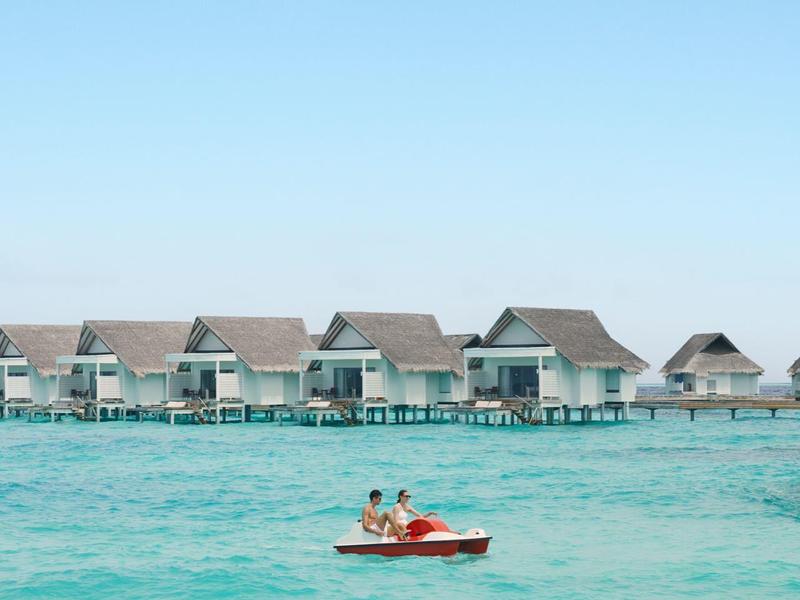 Personen paddeln in einem roten Kajak auf türkisfarbenem Wasser vor Bungalows am Wasser.