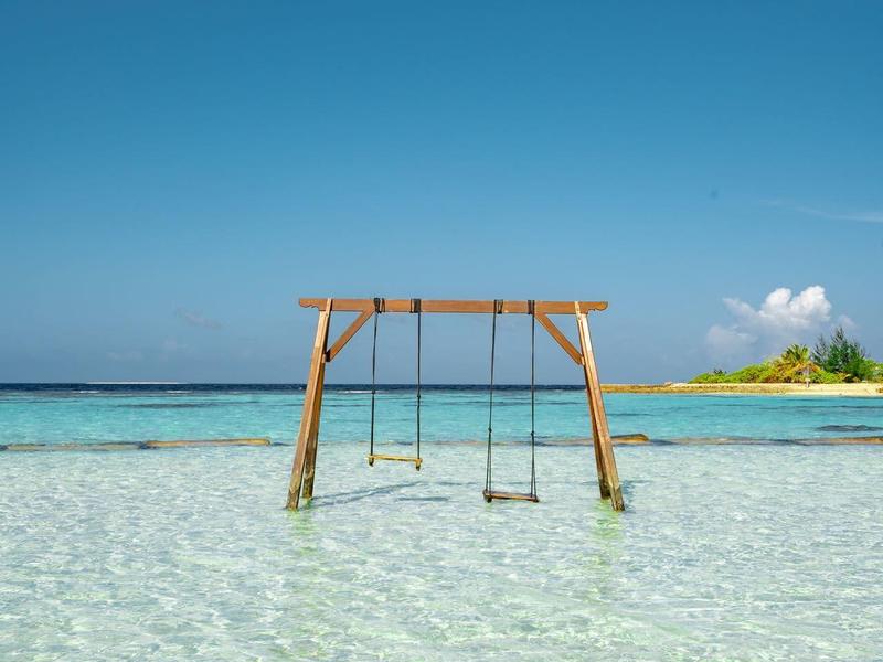 Hölzerschaukel in klarem flachem Meerwasser an einem hellen Sandstrand unter blauem Himmel.