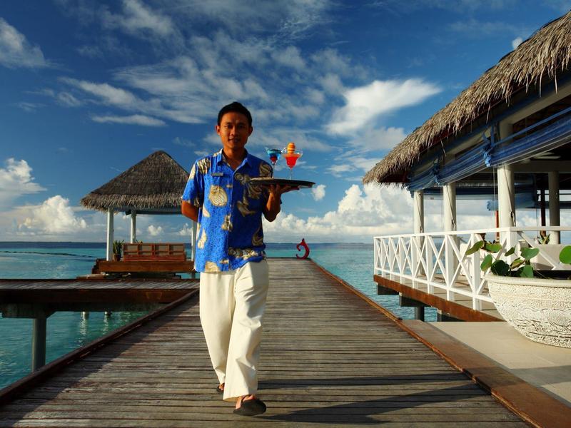 Ein Kellner in blauem Hemd trägt Getränke auf Steg über türkisfarbenem Meer bei Sonnenschein.