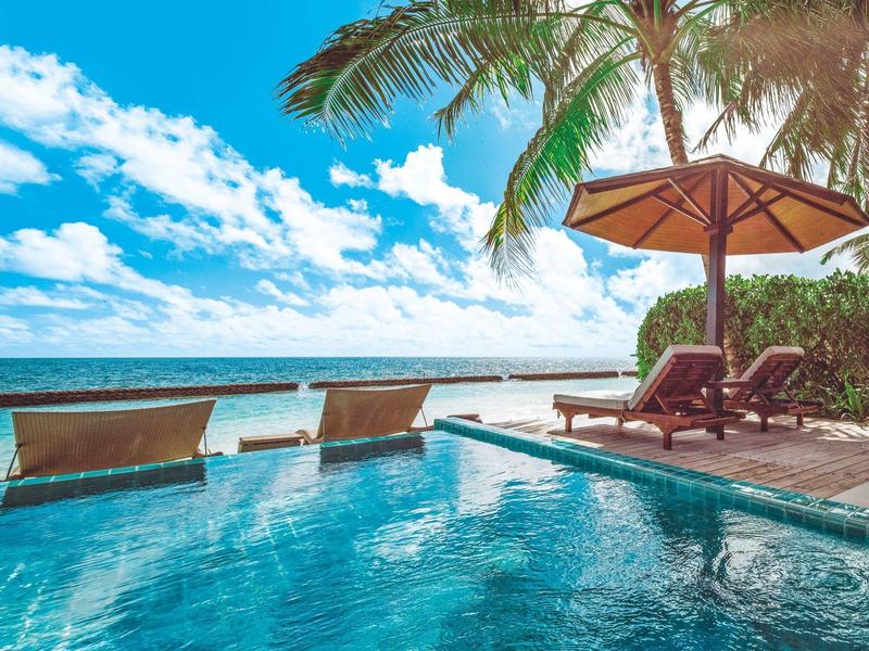 Ein glasklarer Pool mit Liegen und Sonnenschirm, Palmen und blauem Himmel am Meer.