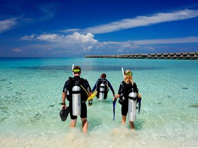Drei Taucher mit Ausrüstung waten im klaren Meerwasser Richtung Steg unter blauem Himmel.