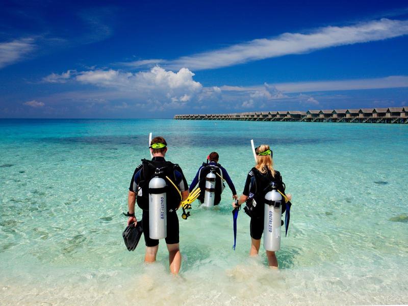 Drei Taucher mit Ausrüstung laufen ins klare Meerwasser an einem sonnigen Strand.