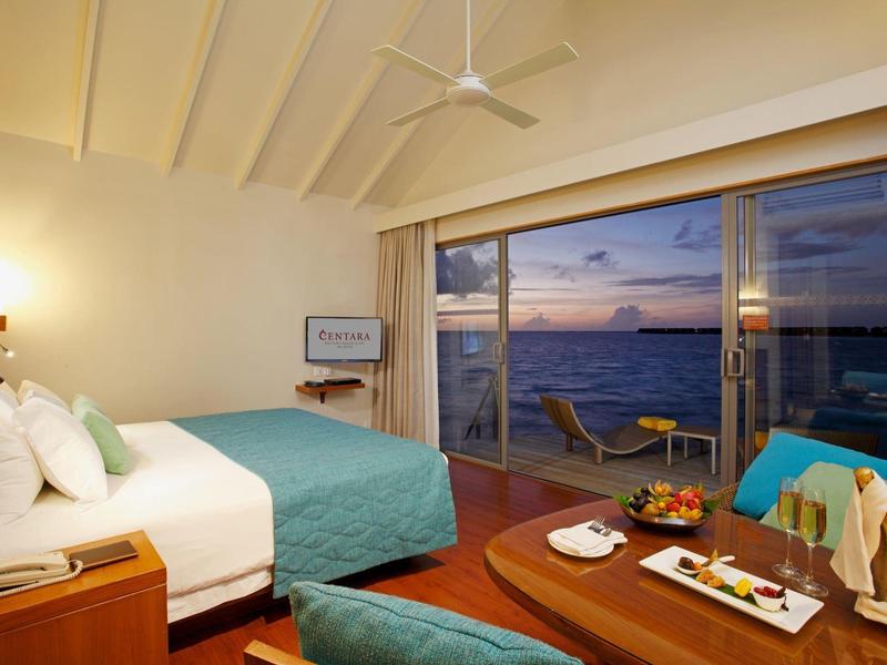 Hotelzimmer mit großem Bett und Blick auf das Meer durch eine Glasfront bei Sonnenuntergang.