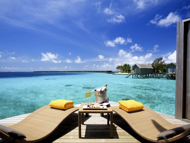 Blick auf türkisfarbenes Meer von zwei Liegestühlen mit Kissen und Tisch dazwischen auf Holzdeck.