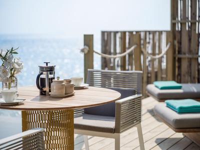 Terrasse mit Holztisch, Stühlen und Liegen mit Blick auf das Meer bei klarem Himmel.