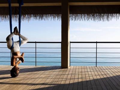 Person macht Yoga-Übung in Hängematte mit Meerblick unter einem Dach mit Holzterrasse.