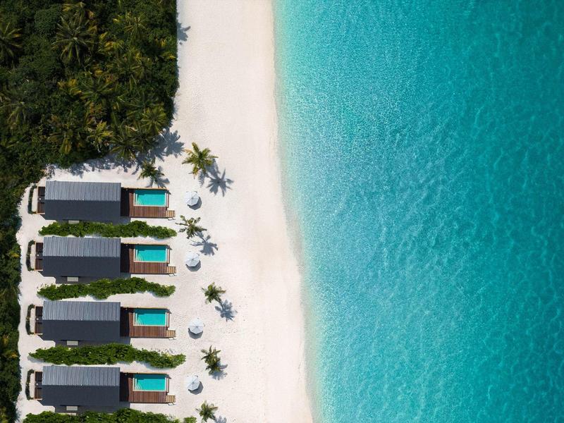 Fünf Villen mit Pools am weißen Strand neben türkisfarbenem Wasser und Palmen.