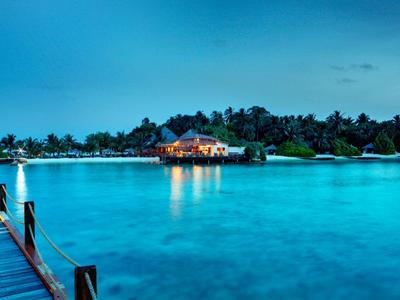 Ein beleuchtetes Haus auf einer Insel, umgeben von blauem Wasser und Palmen bei Dämmerung.